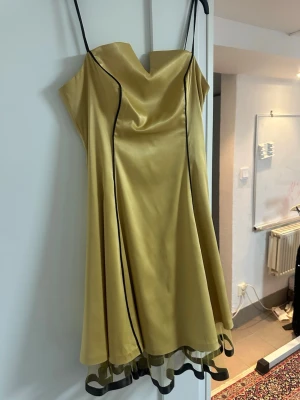 Gul satinklänning med svarta detaljer - Snygg gul klänning i satin med tunna axelband och svarta kantdetaljer. Klänningen har en vacker draperad urringning och volangkant nertill med svart mesh. Perfekt för dig som vill sticka ut med en unik look.