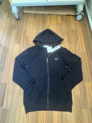 Comme des Garçons - Play hoodie - Svart hoodie med dragkedja från Comme des Garçons Play. Har den ikoniska hjärtloggan med ögon på bröstet och ett stort hjärta med ögon på ryggen. Tillverkad i mjukt bomullsmaterial och har två fickor framtill samt huva med snören.