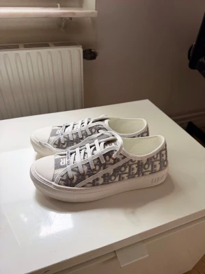 Dior  sneakers - Snygga låga sneakers från Christian Dior i vitt och grått med klassiskt Dior-monogram. Skorna har vita snören, gummisula och textdetaljer på plösen. Perfekta för dig som vill ha en exklusiv och trendig look.