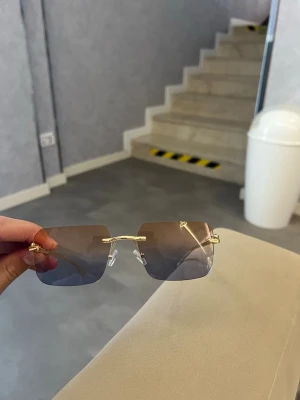 Fyrkantiga solglasögon med gulddetaljer - Säljer ett par stilrena solglasögon med fyrkantiga, tonade glas och tunna guldfärgade bågar. Glasen är utan ram och har en snygg gradient från mörk till ljus. Sidorna har dekorativa detaljer i guld som ger en exklusiv känsla.