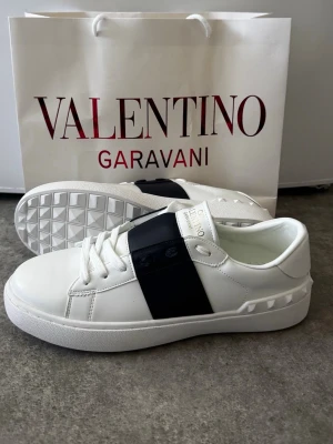 Valentino Garavani Opens - Snygga vita sneakers från Valentino Garavani med bred svart läderdetalj över mitten. Skorna har klassisk låg profil, vita snören och en unik mönstrad sula med nitar längs hälen. Perfekt för dig som vill ha en exklusiv och stilren look.