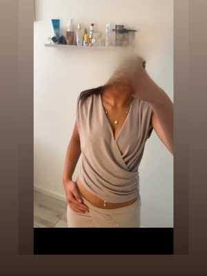 Beige omlottopp med drapering - Snygg beige omlottopp med draperad front och v-ringning. Toppen är ärmlös och har en mjuk, stretchig känsla som gör den bekväm att bära. Perfekt att styla med jeans eller kjol för en trendig look.