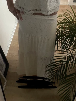 Vit långkjol i bomull med spetsdetaljer - Romantisk vit långkjol i mjuk bomull med fina spetsdetaljer och broderade hålmönster. Kjolen har en avslappnad passform och faller snyggt längs benen. Perfekt för sommarens alla tillfällen.