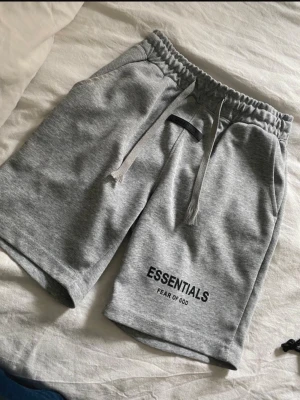 Grå Essentials Fear of God shorts - Snygga grå shorts från Essentials Fear of God med elastisk midja och snörning. Mjuk bomullskänsla och tryckt logga på benet. Perfekta för chill eller streetwear-stil.