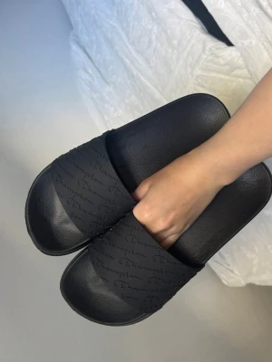 Svarta slides från Champion - Säljer ett par svarta slides från Champion med präglad logga över hela remmen. Sandalerna är gjorda i syntetmaterial och har en klassisk, sportig design med bred rem och bekväm fotbädd. Perfekta för chill dagar eller till stranden.