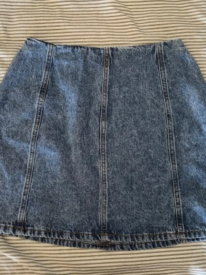 Blå jeanskjol i klassisk modell - Säljer en blå jeanskjol i klassisk kort modell med synliga sömmar framtill. Kjolen har en rak passform och är tillverkad i kraftigt denimtyg. Kjolen är från Carin Wester och är oanvänd. Perfekt att styla med sneakers eller boots för en cool vardagslook.