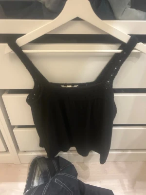 Svart ärmlös linnetopp från H&M - Snygg svart linnetopp från H&M med breda axelband och håldetaljer. Toppen är ärmlös och har en luftig passform, perfekt för varma dagar. Enkel att matcha med både jeans och kjol.