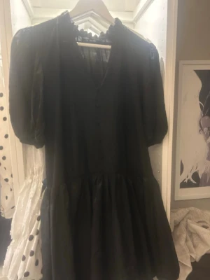 Svart kort klänning från Zara - Snygg svart kort klänning från Zara med puffärmar och volangdetalj nedtill. Klänningen har v-ringning och lätt transparent material upptill. Perfekt för dig som gillar trendiga och feminina plagg.