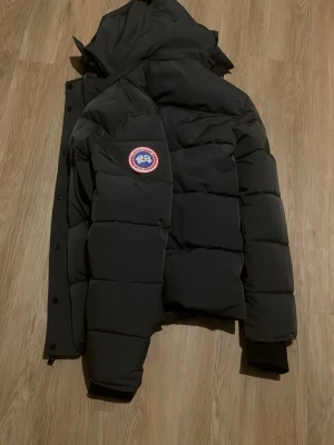 Svart dunjacka från Canada Goose wyndham  - Säljer en svart dunjacka från Canada Goose med klassisk patch på ärmen. Jackan har huva, stora fickor och quiltad design med breda paneler. Perfekt för kalla vinterdagar och riktigt varm tack vare dunfyllningen. Stängs med dragkedja och knappar.