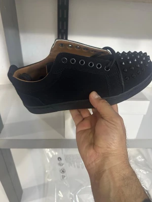 Svarta Louboutin sneakers med nitar - Svarta sneakers från Christian Louboutin med mattsvarta nitar på tån och svarta metalldetaljer. Skorna har en låg siluett, svart mocka och läderfoder. Snygg och edgy design som sticker ut i mängden.