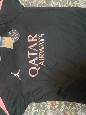 Svart PSG träningströja Jordan och shorts  - Snygg svart träningströja från PSG och Jordan med rosa detaljer och Qatar Airways-tryck på bröstet. Tröjan har rund hals, PSG-logga och Jordan-logga framtill. Perfekt för fotbollsträning eller som sportig streetwear.