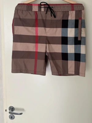 Burberry shorts oanvända  - Säljer ett par klassiska rutig shorts från Burberry med beige, bruna, svarta och röda toner. Shortsen har elastisk midja med svart snörning och sidoficka. Perfekta för sommaren och har den ikoniska Burberry-looken.