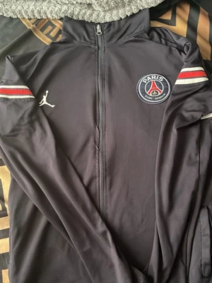 Svart PSG x Jordan träningsjacka - Snygg svart träningsjacka från PSG x Jordan med hel dragkedja, hög krage och broderade loggor på bröstet. Rödvita detaljer på ärmarna och mjukt, stretchigt material som passar perfekt till sport eller chill. Ikonisk Jumpman-logga och PSG-emblem på framsidan.