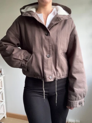 Brun vindjacka med huva och teddyfoder - Snygg brun vindjacka med luva och vitt teddyfoder. Jackan har knäppning med stora knappar, dragsko i midjan och två snedställda fickor. Oversized passform och muddar med knapp vid ärmslut. Perfekt för kyliga dagar.