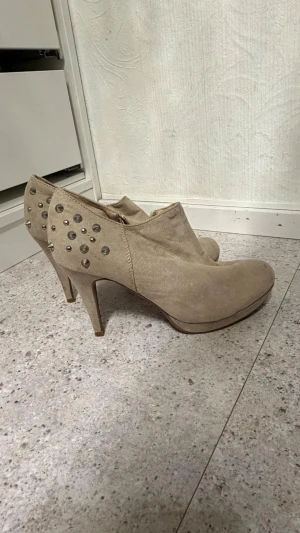 Beige stövletter med nitar - Snygga beige stövletter med spetsig tå och höga smala klackar. Baksidan är dekorerad med silverfärgade nitar för en edgy look. Dragkedja på insidan för enkel påtagning. Materialet ser ut som mocka och ger en mjuk känsla.