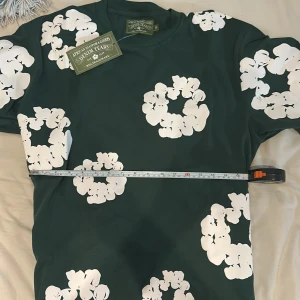 Grön blommig t-shirt Denim Tears - Snygg mörkgrön t-shirt från Denim Tears med stora vita blommiga tryck över hela tröjan. Klassisk passform med rund hals och korta ärmar. Perfekt statement-plagg för dig som vill sticka ut. Materialet är mjuk bomull som känns skönt mot huden. Säljer pågrund avd att den ejj passar mig (för stor) 