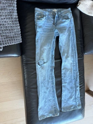 Ljusblå bootcut jeans med slitning - Säljer ett par ljusblå bootcut jeans med slitning på vänster lår och klassiska fem fickor. Jeansen har låg midja och är tillverkade i denim med en lätt tvättad look. Perfekta för en avslappnad och trendig stil.