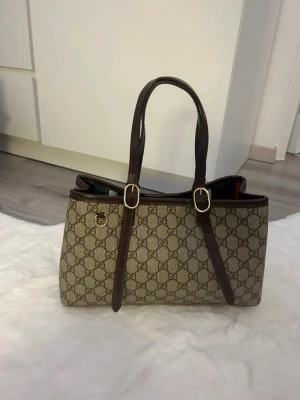 beige monogram handväska - Snygg beige handväska med klassiskt GG-monogram över hela väskan och mörkbruna läderdetaljer. Väskan har två långa handtag med guldfärgade spännen och en stilren, rektangulär form. Perfekt för dig som gillar lyxiga accessoarer.