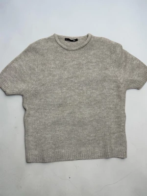 Grå stickad t-shirt från Lager 157 – stl L - Kortärmad stickad t-shirt i ljusgrått från COS. Tröjan har rund halsringning och ribbade kanter vid ärmslut och nederkant. Tillverkad i mjukt material som ger en avslappnad men stilren look.