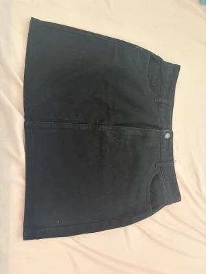 Svart kortkjol i denim - Snygg svart kortkjol i klassisk denim med fickor både fram och bak samt hällor för bälte. Kjolen har en enkel design som passar till många olika stilar och är lätt att matcha med andra plagg.