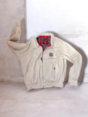 Beige vindjacka från Independent - Stilren beige vindjacka från Independent med röd- och svartmönstrat rutigt foder. Jackan har dragkedja framtill, ribbade muddar och midja samt två sidofickor. Märkets logga finns broderad på bröstet och i nacken. Mudden är lite nopprig men annars är den i bra skick. Storlek S.