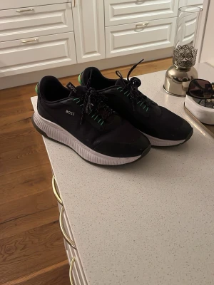 Svarta sneakers från BOSS - Snygga svarta sneakers från BOSS med vita sulor och gröna detaljer på hälen och snörningen. Skorna har en sportig design med rund tå och märkets logga på sidan. Perfekta för en trendig och avslappnad stil. Pris går att diskutera!