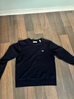 Gant tröja - En mörkblå långärmad tröja med rund halsringning och en liten broderad detalj på bröstet. Tröjan har ribbade muddar vid ärmslut och nederkant. Perfekt för en stilren och enkel look.