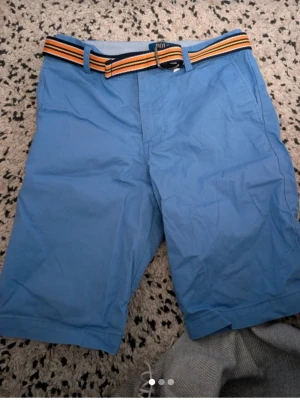 Blå shorts från Polo Ralph Lauren stl 18 (13år 164) - Snygga blå shorts från Polo Ralph Lauren med en randig bälte i orange, svart och gul. Klassisk passform med fickor i sidorna och bälteshällor. Perfekta för varma dagar och enkel att matcha med olika toppar.