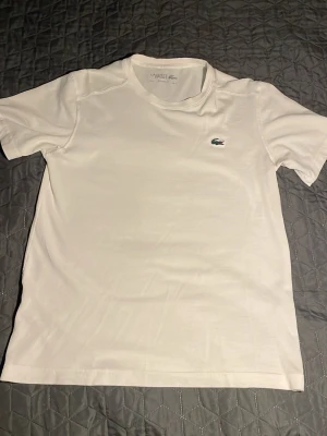 Vit t-shirt från Lacoste - Vit t-shirt från Lacoste med klassisk rund hals och det ikoniska krokodilmärket broderat på bröstet. T-shirten är kortärmad och gjord i mjuk bomull, perfekt för en clean och stilren look. Jätte bra för sommaren som kommer och är bra för att träna eller bara casual fit