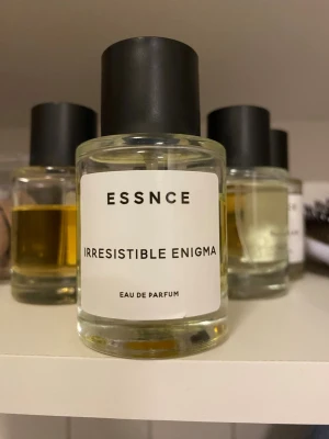 ESSNCE Irresistible Enigma parfym - ESSNCE Irresistible Enigma Eau de Parfum i en stilren, rund glasflaska med svart lock. Parfymen har en ljusgul ton och minimalistisk etikett med svart text. Perfekt för dig som gillar moderna och enkla designer.