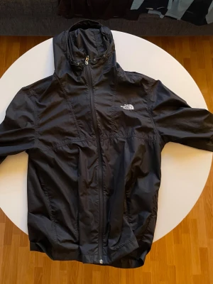 Svart vindjacka från The North Face - Svart vindjacka från The North Face med huva och hel dragkedja framtill. Jackan har justerbar dragsko i huvan och är tillverkad i lätt, vindtätt material. Perfekt för blåsiga dagar och enkel att packa ner i väskan.