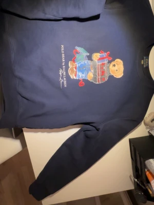 Mörkblå Polo Bear sweatshirt Äkta - Mörkblå sweatshirt från Ralph Lauren med ikoniska Polo Bear-trycket framtill. Björnen har på sig en julig tröja och håller i presenter. Tröjan har rund halsringning och långa ärmar. Perfekt för dig som gillar klassisk och lekfull stil.