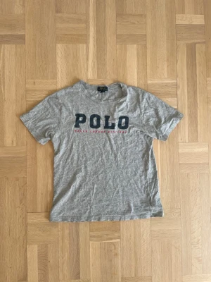 Ralph Lauren T-shirt  - Säljer en grå t-shirt från Polo Ralph Lauren med stort tryck på bröstet. T-shirten är i strl L barn som på bilden och passar enligt lappen 14-16 år. Hör gärna av er om ni har några frågor! MVH tom.