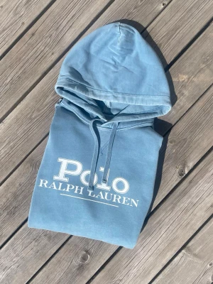 Blå hoodie från Polo Ralph Lauren - Snygg ljusblå hoodie från Polo Ralph Lauren med stor vit logga på bröstet. Klassisk modell med huva, dragsko och känguruficka. Perfekt för en avslappnad och trendig stil.