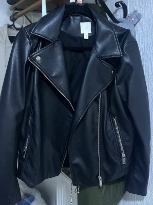 Svart skinnjacka från H&M - Snygg svart skinnjacka från H&M med klassisk bikerstil, stora slag och silvriga dragkedjor. Jackan har två snedställda fickor med dragkedja och en cool, tidlös look. Perfekt för att lyfta vilken outfit som helst.