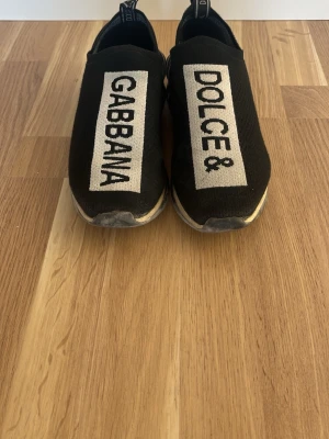 Dolce & Gabbana svarta slip-on sneakers - Svarta slip-on sneakers från Dolce & Gabbana med tydlig logga i vitt på ovandelen. Skorna har en sportig design med elastisk textil och vit sula. Perfekta för dig som gillar streetstyle och vill sticka ut med ett exklusivt märke.