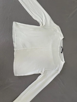 Vit långärmad topp från Zara - Snygg vit långärmad topp från Zara med synliga sömmar och rund halsringning. Tillverkad i mjukt material som känns skönt mot huden. Perfekt att styla med jeans eller kjol🤍