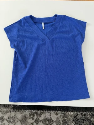 Blå v-ringad topp - En stilren blå topp med v-ringning och korta ärmar. Toppen har en lätt strukturerad yta och en avslappnad passform, perfekt att matcha med jeans eller kjol. Enkel och snygg design utan synliga detaljer eller mönster.