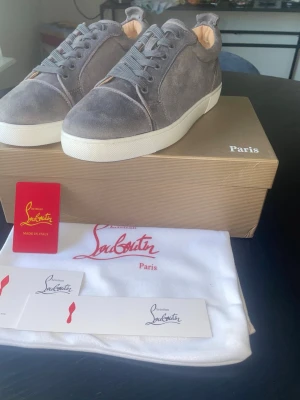 Gråa Christian Louboutin sneakers - Snygga gråa sneakers från Christian Louboutin i mocka med klassisk röd sula. Skorna har lågt skaft, rund tå och vita gummisulor. Kommer med originalkartong, dustbag och certifikat. Perfekta för dig som vill sticka ut med exklusiva detaljer.