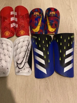 Fyrpack benskydd Nike & Adidas - Säljer fyra par benskydd för fotboll från Nike och Adidas. Ett rött par med Manchester United-logga, ett blått/rött par med FC Barcelona och gul Nike-swoosh, ett vitt par med svart Nike-logga och ett blått/svart par Adidas Predator. Alla har vadderad insida för extra komfort.