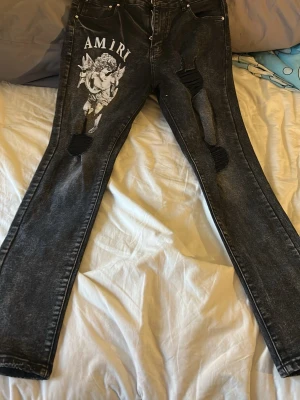 Svarta Amiri jeans - Säljer ett par svarta jeans från Amiri med coolt vitt ängeltryck på vänster lår och slitna detaljer på båda benen. Jeansen har en tvättad look och raka ben. Perfekta för dig som vill sticka ut med streetwear vibe.