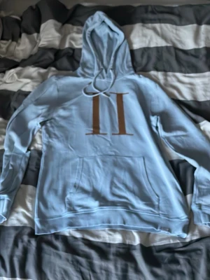 Ljusblå hoodie med brunt tryck - Snygg ljusblå hoodie med stor huva och dragsko. Framtill finns ett brunt tryck med två vertikala streck och en klassisk magficka. Hoodien har ribbade muddar vid ärmslut och nederkant. Perfekt för en chill och avslappnad stil.