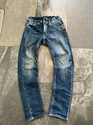 Blå slitna jeans från Replay jeans - Säljer ett par klassiska blå jeans från Replay Jeans med snygga slitningar och tvättade detaljer. Jeansen har fem fickor, raka ben och kontrastsömmar. Perfekta för en avslappnad och trendig look.