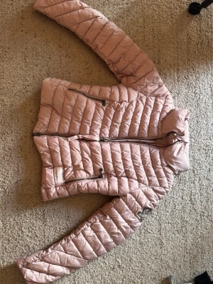 Ljusrosa pufferjacka med dragkedjor - Ljusrosa pufferjacka med två snedställda dragkedjefickor. Jackan har quiltad design och stängs med dragkedja framtill. Perfekt för kalla dagar och har en modern, figurnära passform. Materialet är lätt och ger bra värme.