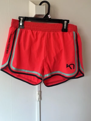 Kari Traa sportiga rosa shorts - Snygga rosa sportshorts från Kari Traa med elastisk midja och kontrasterande turkosa och mörkblå detaljer längs kanterna. Perfekta för träning eller en aktiv dag. Lätta och luftiga i syntetmaterial för maximal komfort. Innershorts 