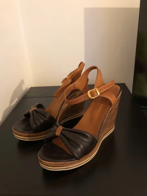 Bruna kilklackade sandaler i äkta skinn - Snygga sandaler med kilklack i trälook, brunt skinn och remmar med guldfärgat spänne. Framsidan har en knutdetalj och sandalerna har öppen tå. Perfekta för sommarens alla outfits.
