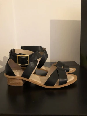 Svarta sandaler med bred klack Clarks - Stilrena svarta sandaler med breda korslagda remmar i äkta läder och en låg, stabil klack i trälook. Sandalerna har en stor guldfärgad spänne vid ankeln och öppen tå. Perfekta för en trendig och bekväm sommarstil.