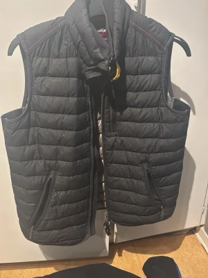Svart dunväst med dragkedjor - Snygg svart dunväst från Parajumpers med quiltad design och två dragkedjefickor framtill. Västen har hög krage med gul spänndetalj och röda sömmar på axlarna. Perfekt för lager-på-lager och streetstyle.