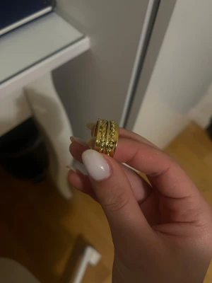 Bred guldfärgad ring med detaljer - Rostfri Snygg bred ring i guldfärg med upphöjda dekorativa detaljer runt om. Ringen har en chunky design och glänsande finish som ger ett statement-intryck. Perfekt accessoar för att lyfta din stil.