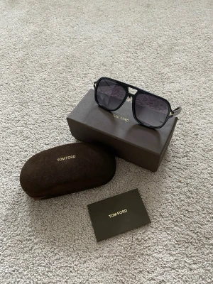 Tom Ford Falconer 02 Mörkblå Faded Linser - Snygga svarta solglasögon från Tom Ford med klassisk pilotform och mörka glas. Bågarna har gulddetaljer vid gångjärnen och kommer med originalfodral och ask. Perfekta för dig som vill ha en stilren och exklusiv look.